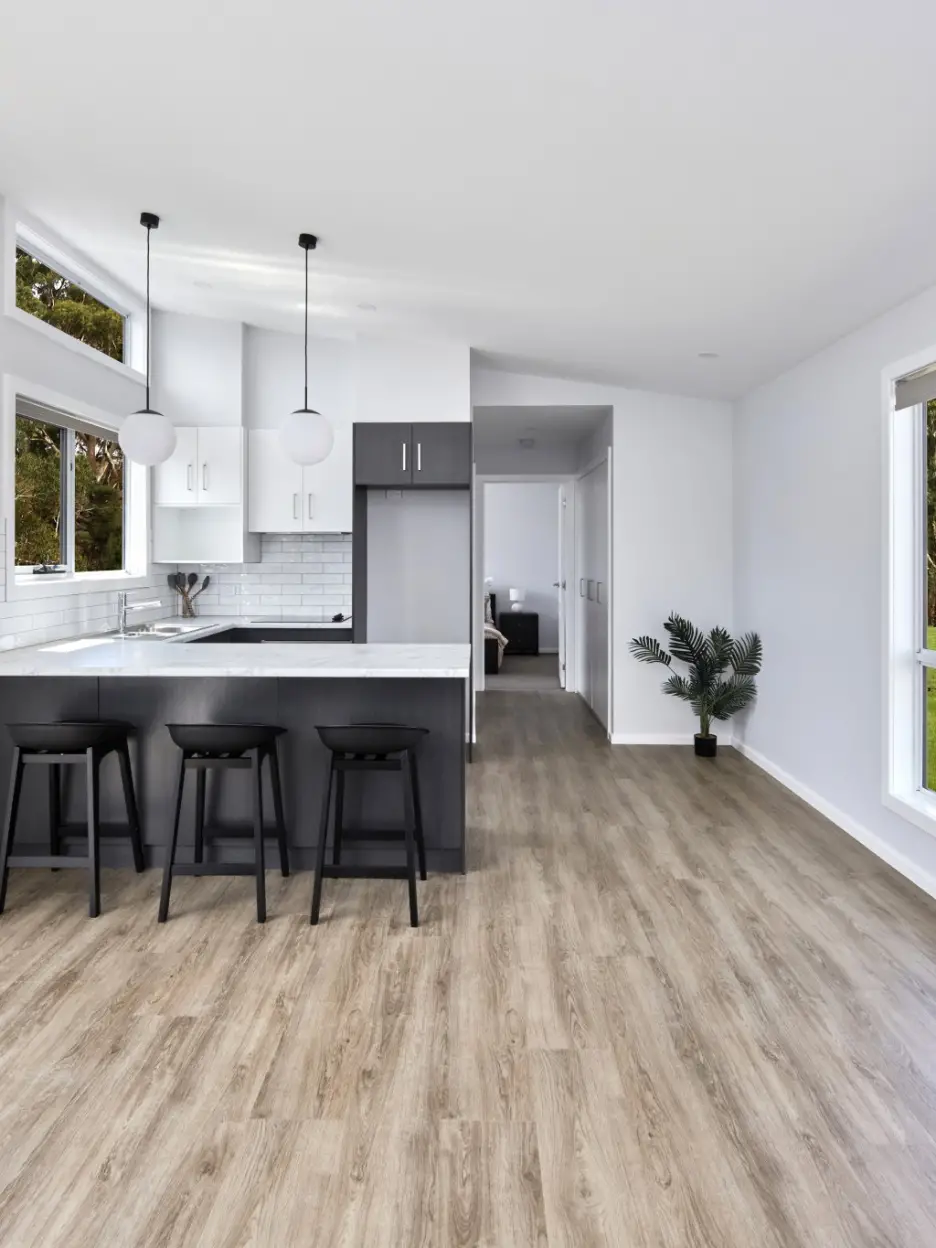 Interior view of the Tasbuilt Homes spec home kitchen, showcasing a white ceiling, walls, benchtop, and tiled splashbacks. The space features dark cabinetry and breakfast bar stools for a modern contrast.