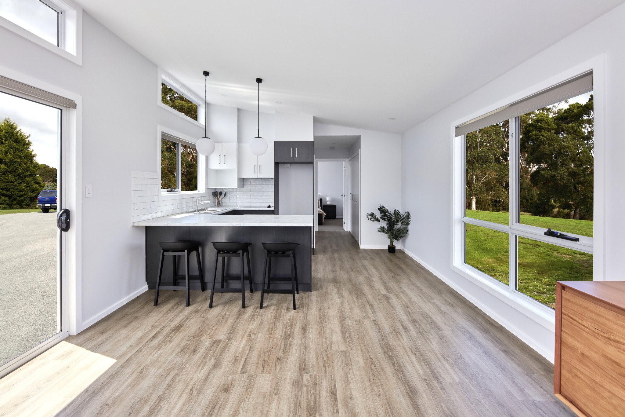 Interior view of the Tasbuilt Homes spec home kitchen, showcasing a white ceiling, walls, benchtop, and tiled splashbacks. The space features dark cabinetry and breakfast bar stools for a modern contrast.
