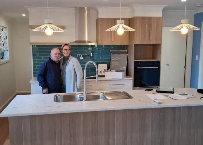 Clients cuddle in their light-filled kitchen with a Tasbuilt gift package. The space includes light timber cabinetry and a green tiled splashback.