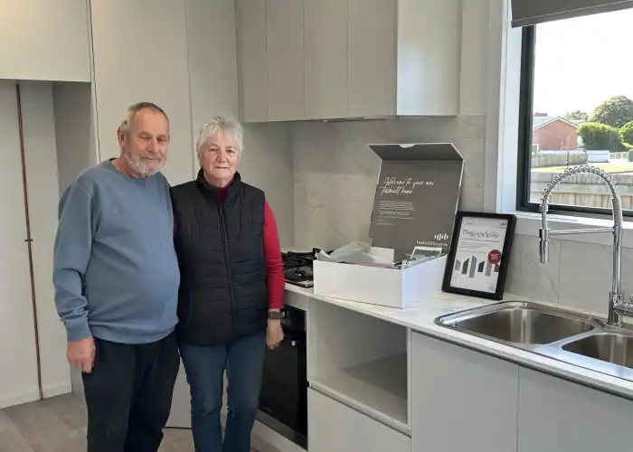 Clients smile in a bright Tasbuilt kitchen with white cabinetry, white benchtops, and silver fixtures.