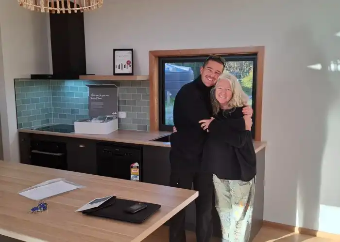 Smiling clients stand in their new Tasbuilt kitchen holding a gift package. The kitchen features warm wooden trims, dark cabinetry, and a green tiled splashback.