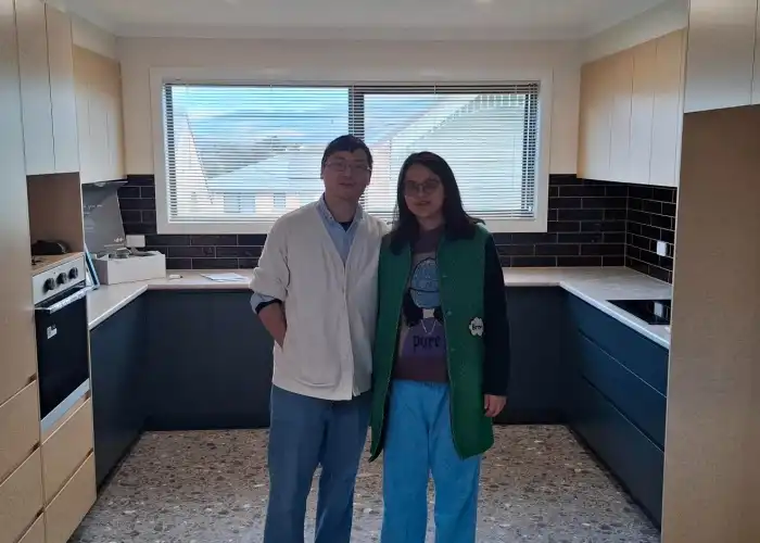 Clients pose in their kitchen with a Tasbuilt gift. The space includes dark blue cabinetry, a blue splashback, and timber features.