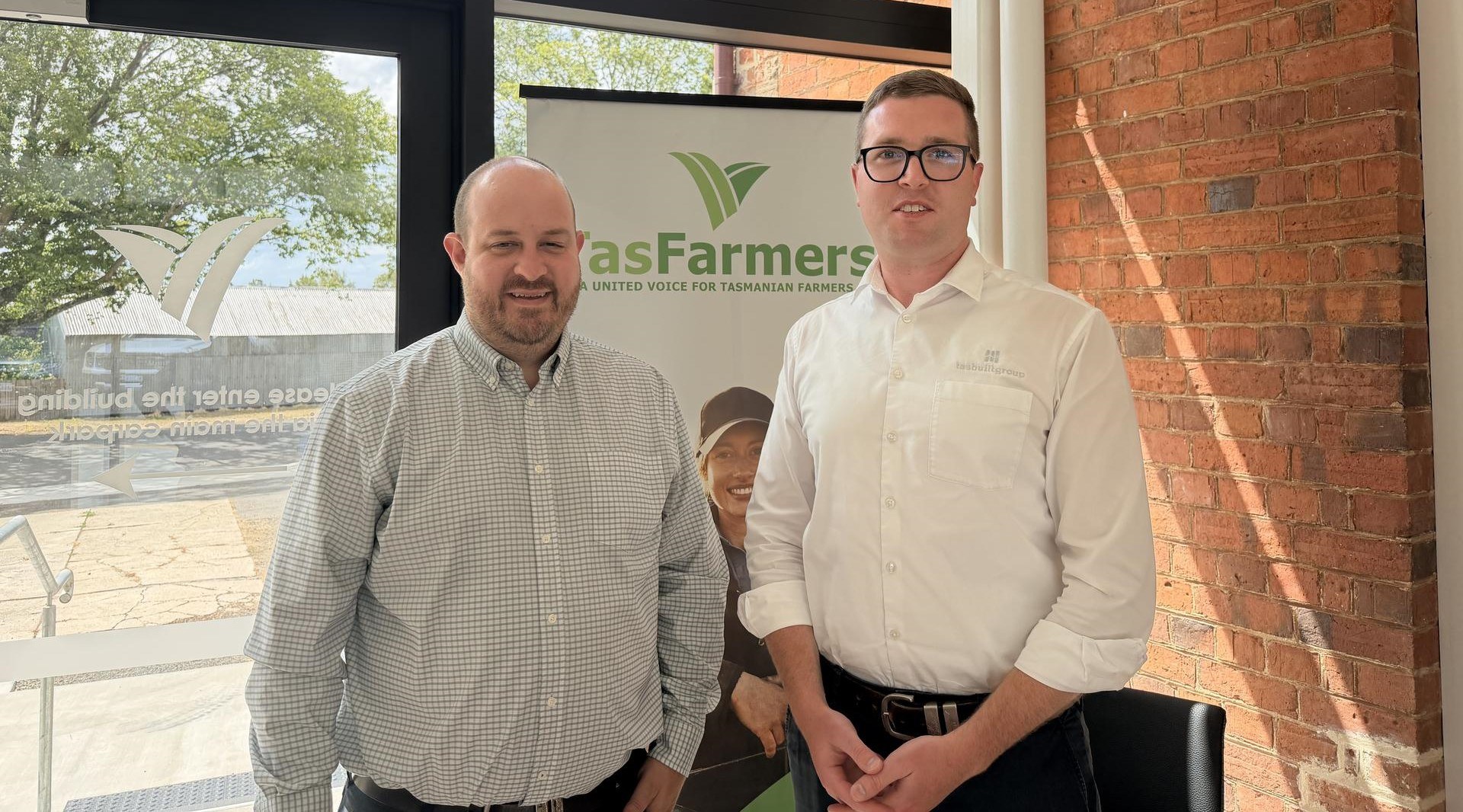 Tasbuilt Commercial and TasFarmers representatives standing in front of a TasFarmers pull-up banner, highlighting their partnership supporting infrastructure for Tasmanian farms.