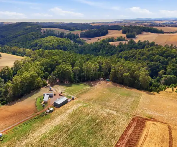 A wide aerial view shows the home situated within a scenic landscape of Tasmanian farmland paddocks and native forests.
